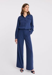 Kobieta stoi z rękami w kieszeniach, nosząc granatową koszulę z długim rękawem zapinaną na guziki oraz szerokie spodnie, a także czarne sandały na obcasach z odkrytymi palcami.