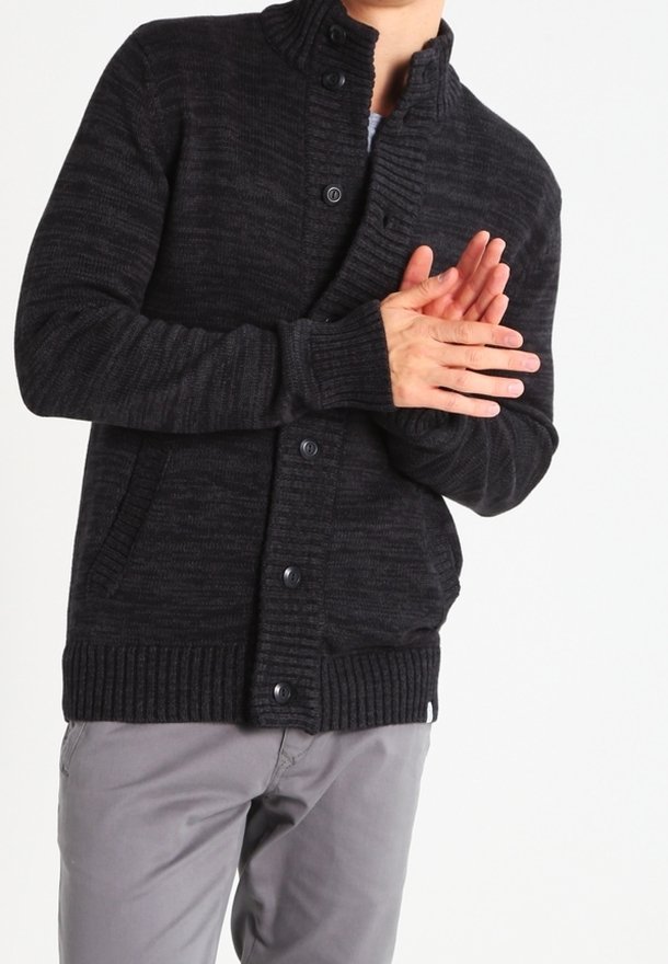 Personne portant un cardigan tricoté foncé boutonné et un pantalon gris clair, les mains jointes devant un fond uni.