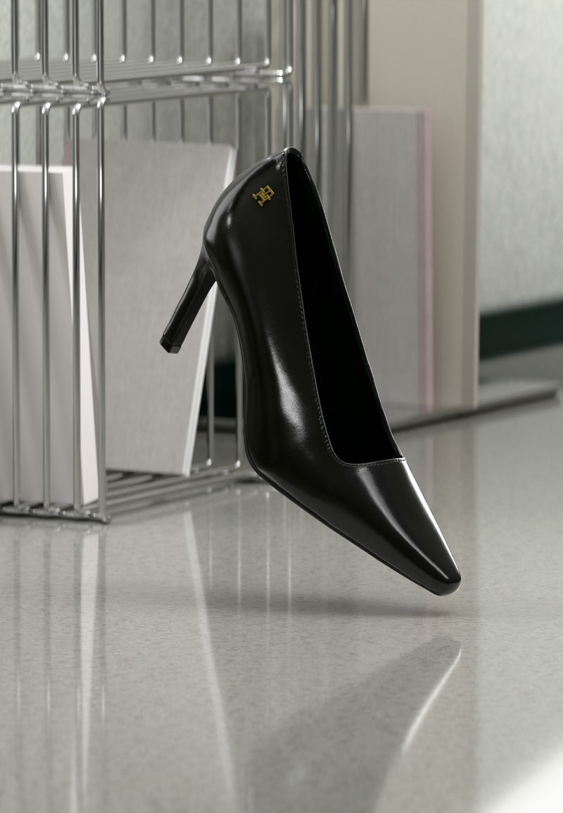 Chaussure à talon haut noire à bout pointu avec un détail logo doré, placée debout sur une surface réfléchissante à côté d'un organiseur de dossiers en métal.