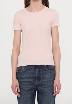 Kobieta ubrana w jasnoróżowy prążkowany t-shirt z krótkim rękawem i ciemnoniebieskie dżinsy, stojąca na tle jednolitego jasnego tła.