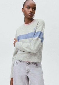 Svetle šedý svetr s modrými pruhy a růžovým límcem, s cropped designem a jemnou texturou, spárovaný se světlými šedými džíny.