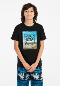 Camiseta negra con un gráfico colorido de una escena de playa que incluye el texto "SOL MAR SURF PROTESTA". Combinada con unos pantalones cortos de baño azules con un patrón de palmeras.