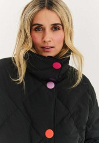 Czarna pikowana kurtka z wysokim kołnierzem, ozdobiona czterema dużymi kolorowymi guzikami w kolorach różowym, fioletowym i pomarańczowym, gładka w dotyku, o minimalistycznym designie.