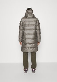 Lang, polstret jakke i grått med hette, har et quiltet design og skinnende tekstur. Brukt over olivenfargede bukser og sneakers.