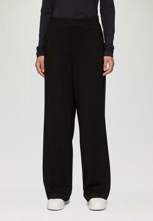 Schwarze weit geschnittene Hose aus strukturiertem Stoff mit hohem Taillenbund, kombiniert mit weißen Sneakers. Das Gesamtdesign ist schlicht und modern.