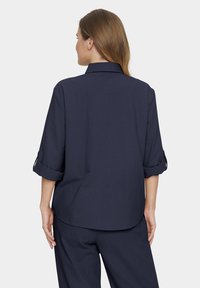 Navy blue, lightweight shirt with rolled sleeves, a classic collar, and a loose fit. Back view showcases a curved hem and smooth texture.