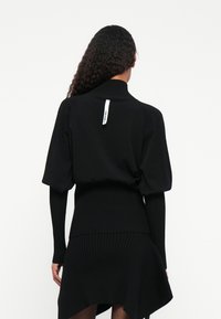 Pulôver preto canelado com gola alta, mangas bufantes curtas e uma bainha traseira única. Apresenta uma etiqueta de marca costurada na gola.