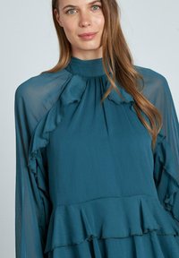 Abito teal con dettagli arricciati, collo alto e maniche lunghe in tessuto trasparente. Tessuto morbido e fluente con un design dell'orlo a strati.