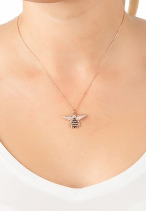 Rose gold halskæde med en bi vedhæng prydet med krystaller, detaljerede vinger og en struktureret krop, som hviler på en hvid bluse.
