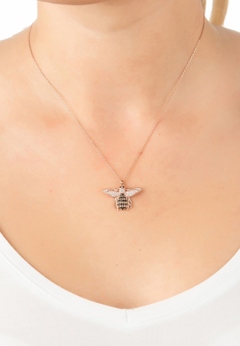 Rózsa arany nyaklánc méhecske medállal, melyet kristályok díszítenek, részletes szárnyakkal és textúrált testtel, egy fehér blúzon pihen.