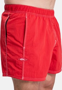 Red swim shorts made of lightweight fabric, featuring elastic waistband, white stitching, side pocket, and logo detail on the left thigh.