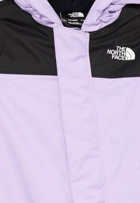 Šviesiai violetinė maža striukė su juodais akcentais, turinti pilną užtrauktuką ir "The North Face" logotipą viršutiniame kairiajame kampe. Lygi audinio tekstūra.