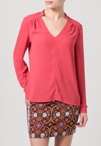 Korálová blůza s dlouhými rukávy a výstřihem do V, spárovaná s vzorovanou mini sukni se květinovými a geometrickými vzory v různých barvách.