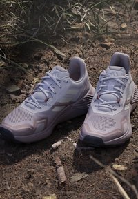 Scarpe sportive grigio chiaro con tessuto in mesh e suole spesse posate su terra asciutta con erba e rami sparsi.