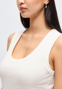 Κοντινό πλάνο μιας γυναίκας με μακριά σκουρόχρωμα μαλλιά, που φορά ασημένια σκουλαρίκια κρίκους και ένα λευκό αμάνικο τοπ με στρογγυλή λαιμόκοψη.