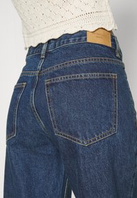 Tmavě modré džínové kalhoty s vysokým pasem, viditelnou světlou koženou značkou na zadní straně a kontrastním žlutým prošíváním na kapsách.