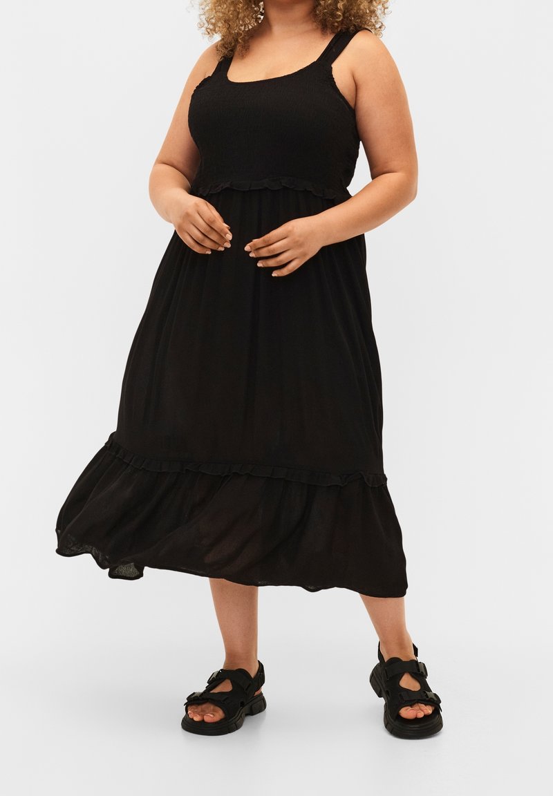 Vestido negro sin mangas con un corsé fruncido, dobladillo con volantes y una silueta fluida. Combinado con sandalias negras con detalles de tiras.