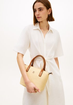 Vrouw in een witte jurk die een beige geweven handtas vasthoudt met bruine leren handvatten en een klein metalen logo aan de voorkant.