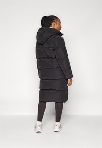 Schwarze puffige Jacke mit Kapuze, mit einem gesteppten Design, langer Silhouette und Reißverschluss. Tragen Sie sie mit dunklen Leggings und Sneakers.