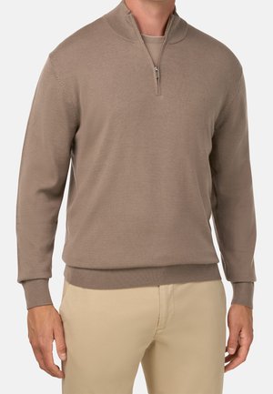 Mand iført beige kvart-glidelås trøje over brune bukser, stående mod en ensartet lys baggrund, hænder afslappet ned langs siderne.
