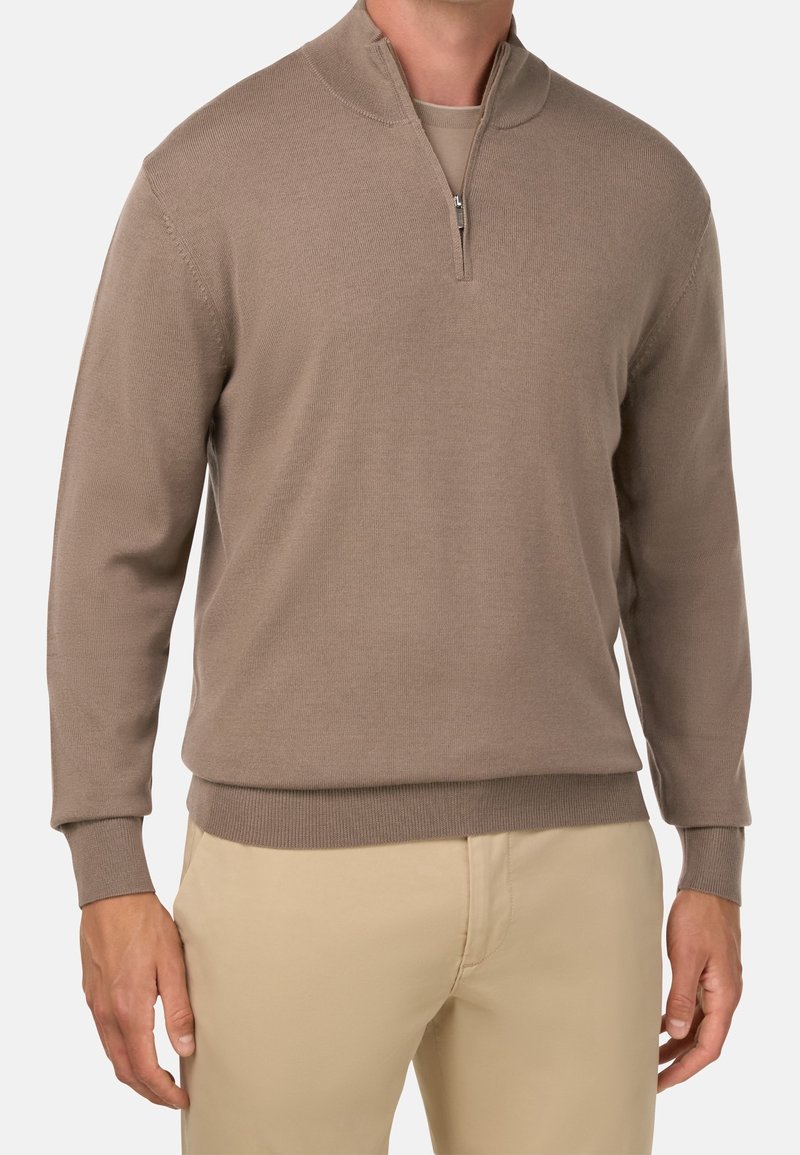 Homme portant un pull beige à fermeture éclair sur un pantalon marron clair, debout devant un fond uni clair, les mains détendues le long du corps.