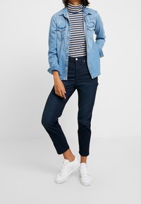 Ljusblå denimjacka över en svart- och vitrandig polotröja, matchad med mörkblå högmidjade jeans och vita sneakers.