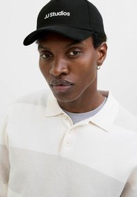 Schwarze Kappe mit "JJ Studios"-Logo, beige gestreiftes Poloshirt mit drei Knöpfen, darüber ein graues T-Shirt. Plain Hintergrund.