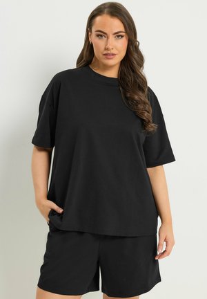 Kobieta z długimi kręconymi włosami, ubrana w luźną czarną koszulkę i pasujące czarne szorty, stoi na białym tle.