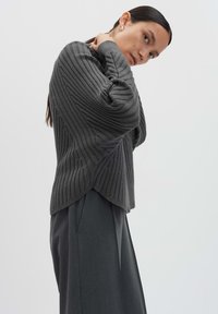 Donna che indossa un pullover a coste grigio scuro con maniche a pipistrello e pantaloni larghi grigio scuro su uno sfondo chiaro e semplice.