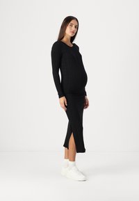 Μαύρο ριχτό φόρεμα εγκυμοσύνης με μακριά μανίκια και πλάγιες σχισμές, με εφαρμοστή σχεδίαση. Συνδυάζεται με λευκά αθλητικά παπούτσια και κάλτσες μέχρι τον αστράγαλο.