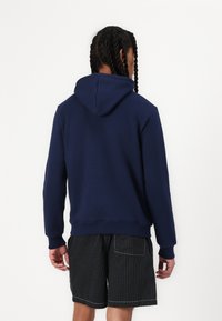 Mørkeblå hettegenser med snorhette, myk stoff, ribbede mansjetter og hem, iført med svarte stripete shorts. Bakfra viser det rene linjer.