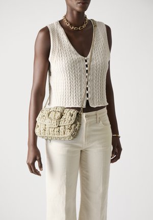 Beige woven handbag with a quilted texture, a gold chain strap, and decorative hardware. Worn crossbody on a neutral outfit.
