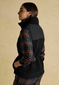 Czarna pikowana kamizelka z wysokim kołnierzem, zestawiona z wielokolorową koszulą w kratę w odcieniach czerwieni, niebieskiego i zielonego. Tekstura jest miękka i puszysta.