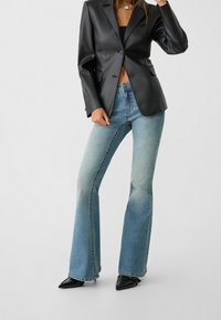 Sort blazer i kunstlæder med to knapper og slagkrave; parret med lyseblå flared jeans i en lysnet tekstur; sorte spidse hæle.
