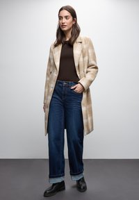 Lungo cappotto beige a quadri sopra un top marrone, abbinato a jeans a gamba larga di colore blu scuro risvoltati alle caviglie e stivaletti neri.