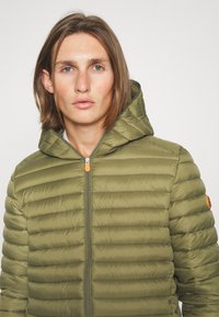Groene pufferjack met verticale stiksels, ritssluiting aan de voorkant en capuchon. Kenmerkt een oranje ritssluiting en een logo-accent op de mouw.
