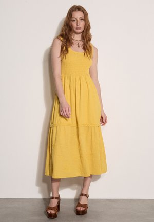Junge Frau mit langen rötlichen Haaren, die ein ärmelloses gelbes Midi-Kleid und braune Plateausandalen trägt, steht vor einer schlichten Wand.