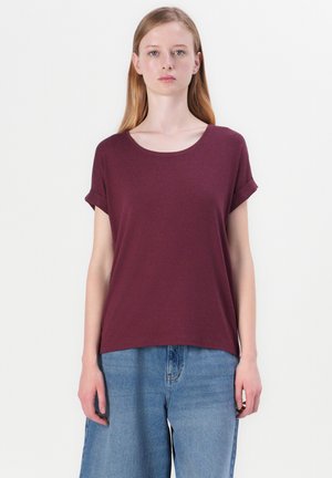 Jeune femme aux cheveux longs et raides, portant un t-shirt bordeaux à manches retroussées et un jean bleu ample, se tenant devant un fond blanc.