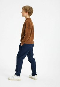 Brązowy sweter robiony na druty z wzorem w kratkę, zestawiony z granatowymi spodniami i białymi sneakerami. Widok z boku, na tle białego tła.