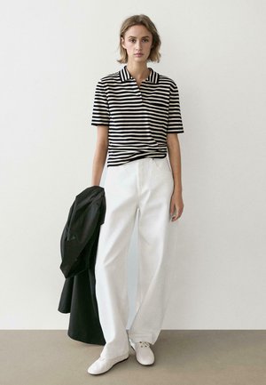 Giovane donna in piedi contro un muro bianco, indossa una maglietta a righe bianche e nere, pantaloni bianchi, scarpe bianche, tiene un cappotto nero.