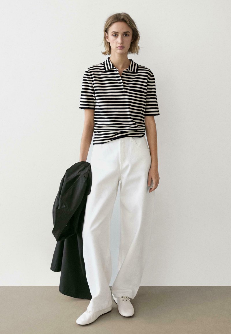 Jeune femme debout contre un mur blanc, portant un chemisier rayé noir et blanc, un pantalon blanc, des chaussures blanches, tenant un manteau noir.