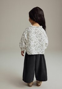 Vit blus med blommönster och volangkrage, kombinerad med svarta vida byxor och beige sandaler, sedd bakifrån.