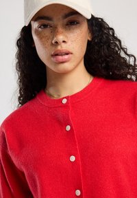 Nuori nainen, jolla on kihara tumma hius, pisamia, beige lippalakki, punainen nappitakki ja ohut kultaketju neutraalilla taustalla.