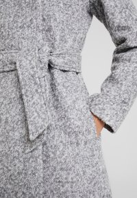 Manteau gris texturé avec une taille ceinturée, des manches longues et des poches latérales, fini avec un tissu doux et duveteux.