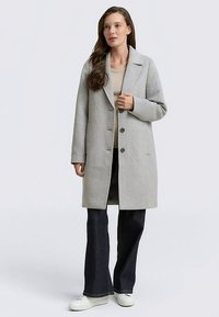 Helehall villasegu mantel lühikeste kraeavadega, kolme esinööbi ja küljetaskutega, lõdva lõikega, kombineeritud tumedate teksade ja valgete tossudega.