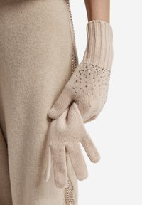 Gants tricotés beiges avec poignets côtelés, présentant un motif ponctué de petits strass sur la paume. Texture douce et forme ajustée.