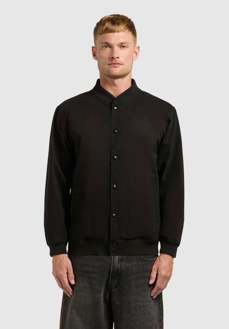 Jeune homme portant une veste bomber noire boutonnée et un jean gris foncé, debout devant un fond uni gris clair.