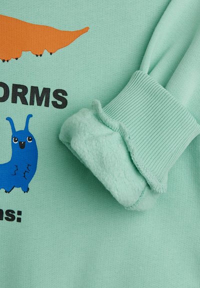 Ανοιχτό πράσινο ύφασμα με μανίκι με περιλαίμιο και πολύχρωμες καρικατούρες κάμπιες με μερικό μαύρο κείμενο.
