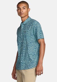 Camisa de botones de manga corta en color verde azulado con estampado de leopardo. Presenta un cuello extendido y un pequeño logotipo bordado en el lado izquierdo del pecho.