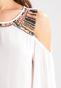 Camicia bianca con design a spalle scoperte, collo decorato con cinturini colorati e dettagli in perline, realizzata in tessuto leggero.